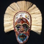 Tribal-style mask with large tan fibers representing hair, a dark brown face. The eyes have circles of black around them and the outside has a circle of blue as if the mask is wearing ceremonial paint.