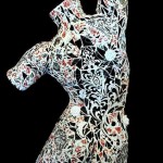 Back view of sculpture of female torso in reds, black, and white in a dancer's position. She has white flowers for nipples and positioned around her body, a large white flower between her legs, and a small red heart under her left breast.