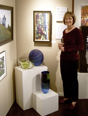 Photograph of Susan Koch standing next to an exhibit of her paintings.