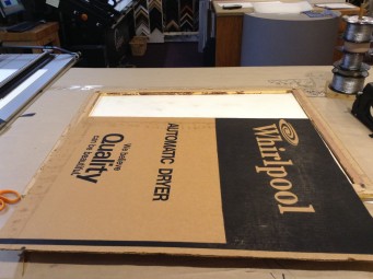 Cardboard cut from appliance box being used as backing for framed artwork.
